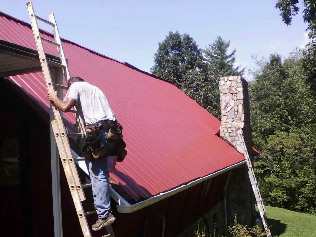 Roofer on a ladder beside a red metal roof and stone chimney — Soffit & Fascia Repair by Roofing Brothers in South Saint Paul